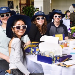 一群面带微笑的年轻人坐在一张桌子旁, 每个人都戴着白色的太阳镜和相配的蓝色帽子. 他们被礼品盒、小册子和插花包围着. 场景似乎是一个非正式的活动或聚会.